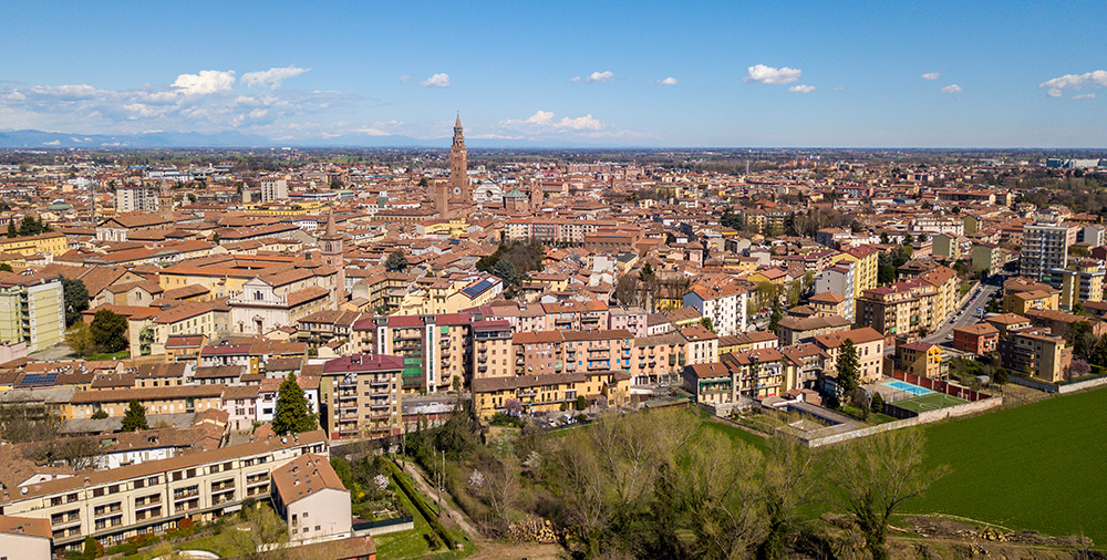 Aerial view of a European town with terracotta rooftops, a tall church bell tower, and surrounding fields under a clear blue sky.